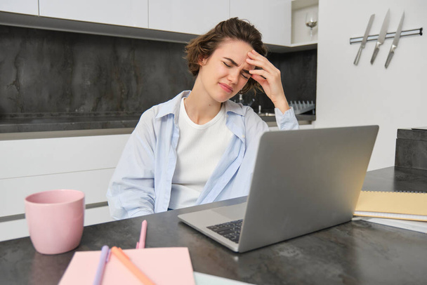 Woman has headache while works from home. Girl has painful migraine, sits near laptop, grimaces from discomfort. - Photo, Image