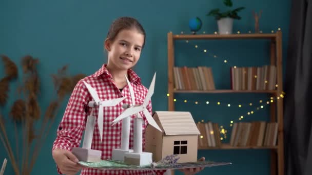 Nastolatka pozująca z modelką turbiny wiatrowej i modelką domu w domu. Przestrzeń kopiowania - Materiał filmowy, wideo
