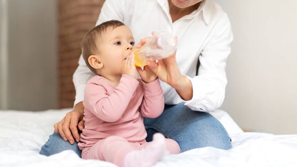 Sede bonito bebê bebê menina bebendo água de garrafa, sentado com sua mãe na cama, close-up, panorama. Criança desfrutando de bebida saudável, cortada - Foto, Imagem