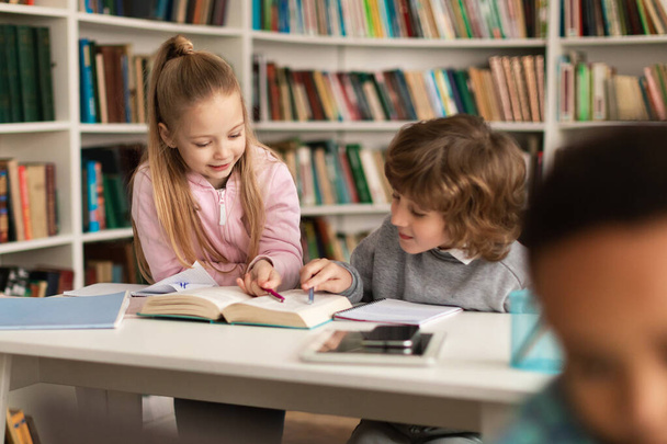 School pupils reading book together in library, doing homework or preparing for classes, sitting at desk and chatting. Education, kid and study concept - Photo, Image