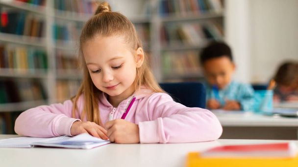 В начальной школе. Портрет довольно умной школьницы, которая пишет в тетради и улыбается, сидит за столом в классе с одноклассниками на заднем плане, панорама, свободное пространство - Фото, изображение