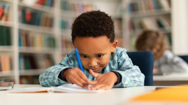 Концентрований чорний школяр сидить за столом у школі і вчиться писати в книзі з олівцем, навчанням, панорамою. Різні діти в класі - Фото, зображення