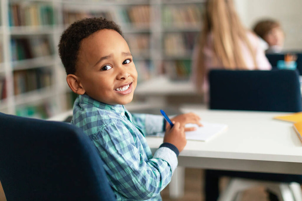 Portré afro-amerikai általános iskolás fiúról, aki az asztalnál ül az osztályteremben, visszafordul és mosolyog a kamerára, szabad tér. Osztálytársak tanul a háttérben - Fotó, kép