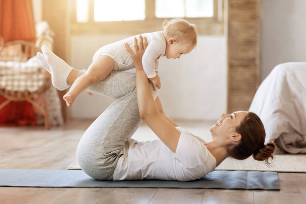 Sport, motherhood and active lifestyle concept. Side view of young mother workout together with her baby toddler in living room at home. Sporty mom having fun and playing with her little infant child - Photo, Image