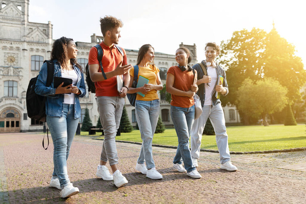 Cinque studenti multietnici che chiacchierano e ridono mentre camminano insieme al campus universitario, un gruppo di giovani felici con libri e zaini che vanno a lezione o si divertono dopo le lezioni - Foto, immagini