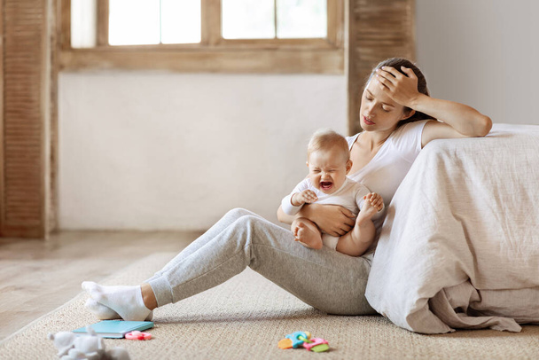 Tired infant baby cries in mother hands, depressed unhappy exhausted mom sitting on floor with crying little child on her lap, bedroom interior, copy space. Postnatal postpartum depression concept - Photo, Image
