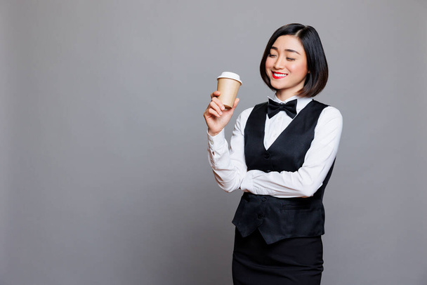 Lächelnde asiatische Kellnerin in Berufskleidung, die Kaffee aus Pappbechern trinkt. Fröhliche junge Empfangsdame steht mit geschlossenen Augen und genießt Heißgetränk to go - Foto, Bild