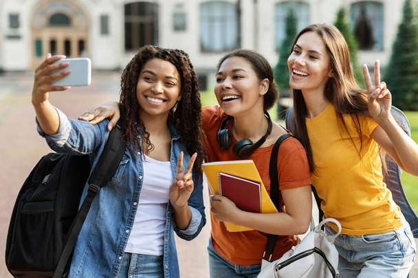 Üç çok ırklı kız öğrenci dışarıda akıllı telefondan selfie çekiyor. Sırt çantaları ve çalışma kitaplarıyla birlikte cep telefonu kameralarına poz verip dersten sonra dinleniyorlar. - Fotoğraf, Görsel
