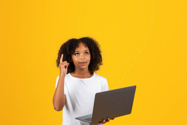Cheerful smart adolescent black girl in white t-shirt, uses laptop for study, got idea, point finger up, isolated on orange background, studio. Device, app for knowledge, modern education at school - Photo, Image