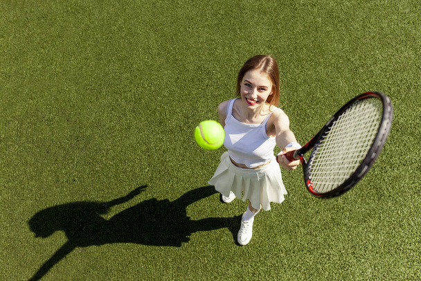 fiatal lány teniszező fehér sport egyenruha teniszezik a zöld pályán, női edző tenisz ütő üti a labdát a levegőben, felső nézet - Fotó, kép