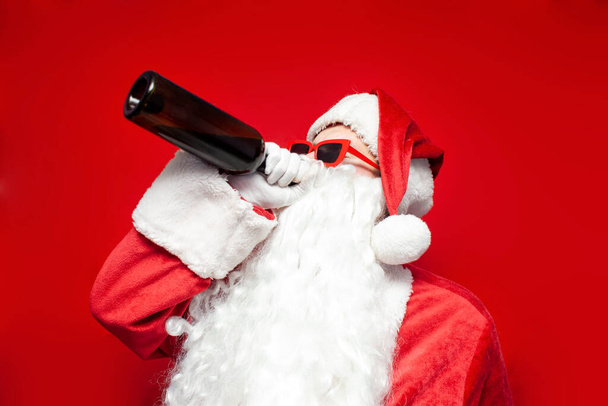 пьяный Санта Клаус в шляпе и праздничные бокалы пьет вино из бутылки на красном фоне, мужчина в костюме Санты пьет алкоголь на Рождество - Фото, изображение