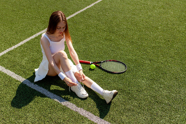 młoda dziewczyna tenisistka w białym mundurze sportowym wiąże sznurowadła na tenisówkach na zielonym korcie, trenerka z rakietą tenisową, przestrzeń do kopiowania - Zdjęcie, obraz