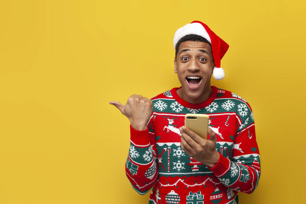 african american guy in christmas clothes and santa hat uses smartphone and shows his hand to the side on yellow background, man in sweater types online on phone and advertises copy space - Photo, Image