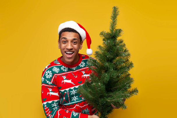 chico afroamericano en ropa de navidad y sombrero de santa sostiene árbol de navidad en bolsa sobre fondo aislado amarillo, hombre en suéter compra decoraciones para el nuevo año - Foto, Imagen