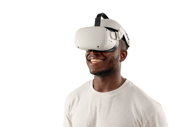 Afrikanisch-amerikanischer Mann in weißem T-Shirt schaut in Virtual-Reality-Brille auf weißem Hintergrund weg und lächelt, Typ mit Vr-Brille schaut in Kopierraum - Foto, Bild