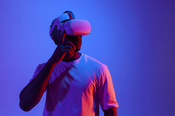 hombre afroamericano jugando videojuego en gafas de realidad virtual en iluminación de neón, el chico utiliza gadget vr moderno y mira hacia otro lado en el espacio de copia - Foto, imagen