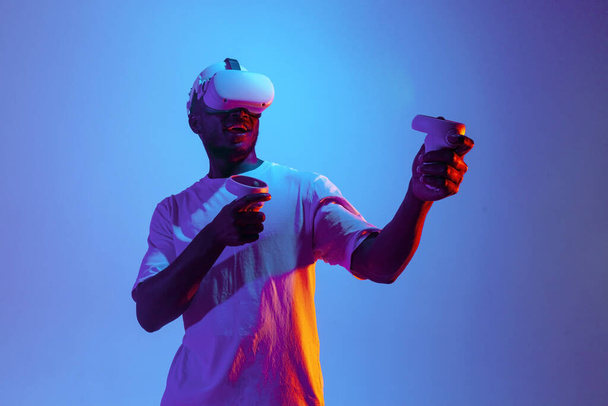choqué gamer homme afro-américain en réalité virtuelle lunettes tire et vise avec joysticks dans l'éclairage au néon, le gars utilise gadget vr moderne et détient panneau de commande - Photo, image
