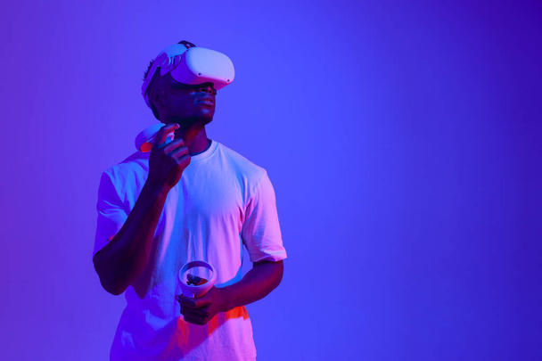 african american male gamer in virtual reality glasses with joysticks looks away in neon lighting, guy plays with vr gadget and holds control panel and looks at copy space - Foto, immagini