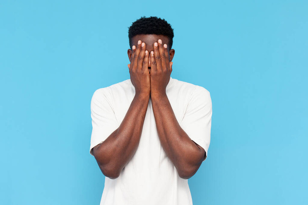 Afrykański Amerykanin w białej koszulce jest nieśmiały i boi się na niebieskim, odizolowanym tle, facet w stresie zakrywa twarz rękami i chowa się - Zdjęcie, obraz