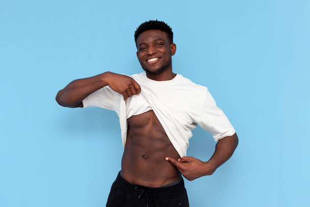 Afrikkalainen amerikkalainen urheilullinen mies näyttää vatsalihasten sininen eristetty tausta, kaveri nostaa t-paita ja näyttää lehdistölle, käsite kunto ja urheilu - Valokuva, kuva