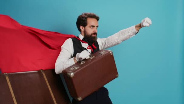 Hotel-Concierge verhält sich wie ein Superheld mit rotem Umhang und bietet an, Menschen mit Koffern vor der Kamera zu helfen. Junger erwachsener Türsteher präsentiert Comicfigur mit Mantel, fühlt sich entschlossen und selbstbewusst. - Filmmaterial, Video
