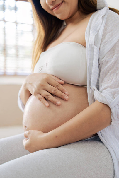 Glückliche weibliche Schwangerschaft sitzt auf dem Sofa und hält ihren Bauch im Wohnzimmer. - Foto, Bild