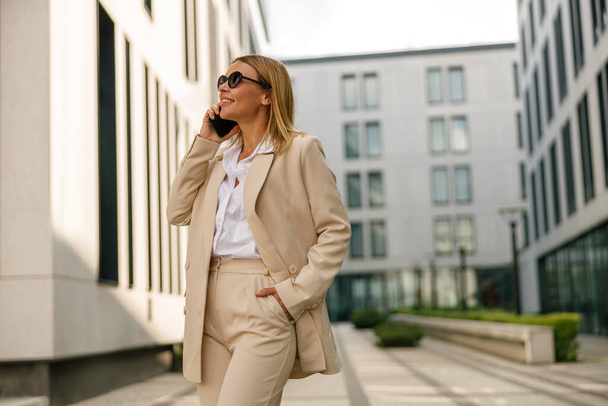 Χαμογελαστή υπάλληλος γραφείου γυναίκα στέκεται στο σύγχρονο κτίριο φόντο και μιλάμε τηλέφωνο - Φωτογραφία, εικόνα