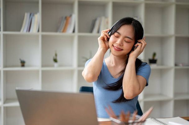 Una encantadora y feliz joven asiática estudiante universitaria disfruta de la música en sus auriculares en su mesa de estudio en casa. Concepto de estilo de vida - Foto, Imagen
