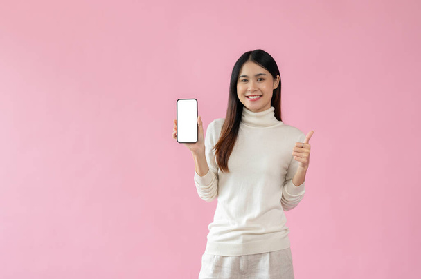 Приваблива і усміхнена азіатка в повсякденному одязі показує великий палець і порожній екран смартфона в руці до камери, стоячи на ізольованому рожевому тлі. - Фото, зображення