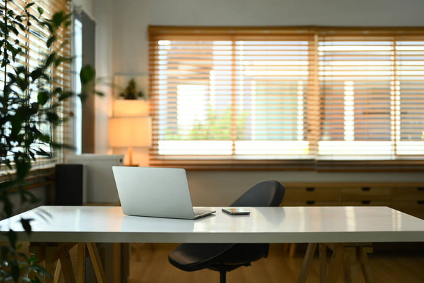 Thuis kantoor interieur met laptop op witte tafel, potplant en warm licht van de lamp. - Foto, afbeelding