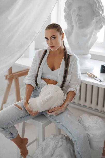 Female sculptor artist captured in her workshop, wearing a loose white shirt and denim jeans with suspenders while holding a historic Greek bust, sitting on a chair amidst her sculptures - Photo, Image