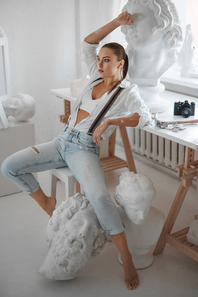 An attractive female sculptor dressed in a loose white shirt and jeans with suspenders, while sitting on a stool in her sculptors studio - Photo, Image