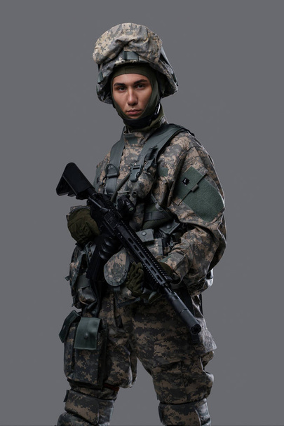 Молодой человек в военной форме и шлеме держит винтовку на сером фоне - Фото, изображение