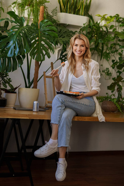 A charming blonde girl in white shirt and denim jeans enjoys sushi sitting on a table surrounded by potted plants - Фото, изображение