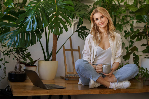 Lovely blonde woman enjoys a cup of tea while sitting next to her laptop on a table surrounded by potted plants - Photo, Image
