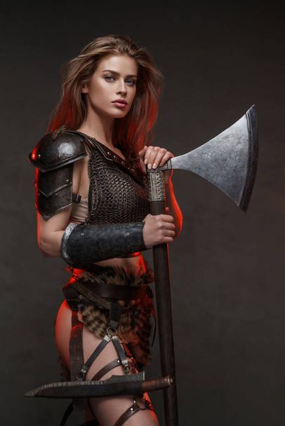 Superbe femme viking vêtue d'un haut de maille et d'une jupe en fourrure avec une hache à deux mains éclairée par un éclairage de bord rouge  - Photo, image