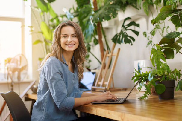 Красивая улыбающаяся внештатная женщина в голубой рубашке работает на ноутбуке в освещенной солнцем комнате, заполненной растениями - Фото, изображение