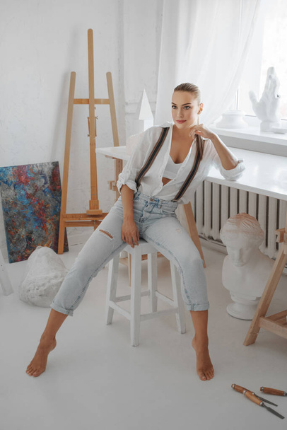 Heykeltraş stüdyosundaki bir taburede otururken beyaz gömlek ve pantolon giymiş çekici bir kadın heykeltıraş. - Fotoğraf, Görsel
