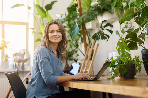 Okouzlující usmívající se žena na volné noze v modré košili pracující na svém notebooku v místnosti plné rostlin - Fotografie, Obrázek