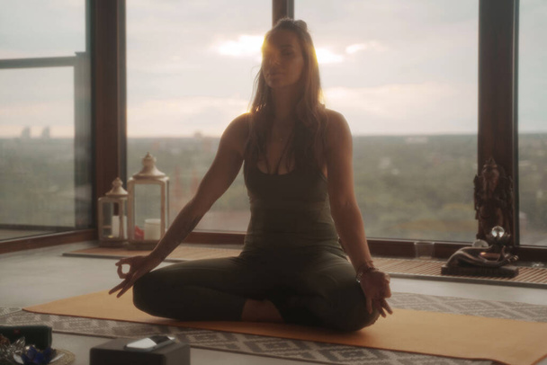 Mulher em trajes de esportes verdes praticando ioga e meditação em pose de lótus contra um cenário panorâmico da cidade em um apartamento - Foto, Imagem