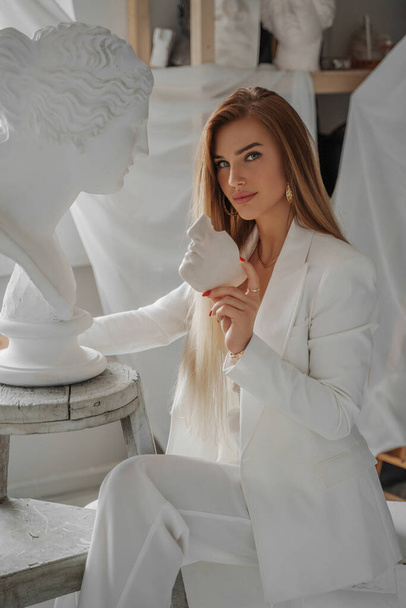 Exquisite young woman, with a models appearance, wearing a stylish white suit and seductive bra top, holding piece of plaster mask while sitting in a sculptors studio near ancient Greek bust - Photo, Image