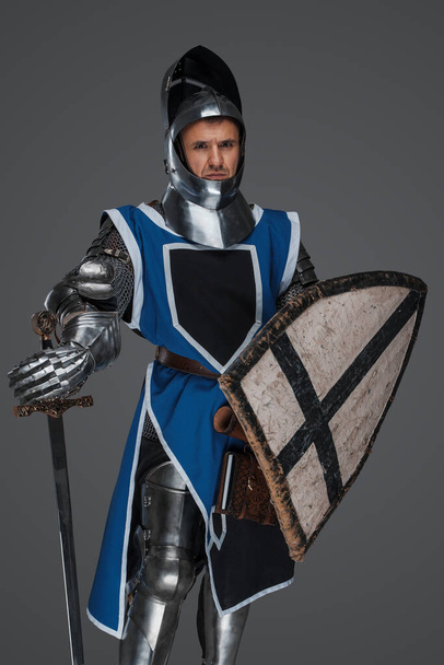 Statečný středověký válečník v brnění a modrém plášti se zvednutým hledím, svírající meč, statečně hledící do kamery, zasazený do šedého pozadí - Fotografie, Obrázek
