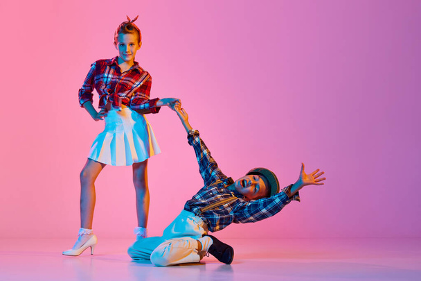 Beautiful children, boy and girl in stylish retro clothes dancing against pink studio background in neon light. Concept of childhood, hobby, active lifestyle, performance, art, fashion - Zdjęcie, obraz