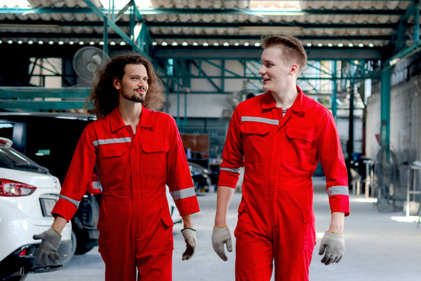 Two handsome mechanic men in red uniform walking through row of vehicle at garage, auto mechanic technician friend work together to repair and maintenance customer car automobile at car service shop. - Foto, imagen