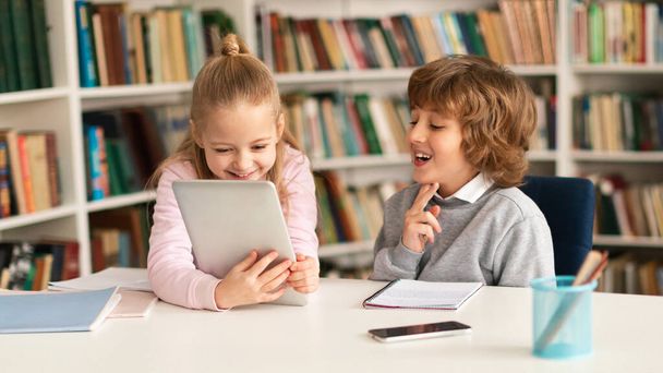 Két általános iskolás gyerek ül együtt az íróasztalnál az osztályteremben vagy a könyvtár belsejében, digitális táblagépet használva, játékokat játszva vagy oktatási videókat nézve a szünetben - Fotó, kép