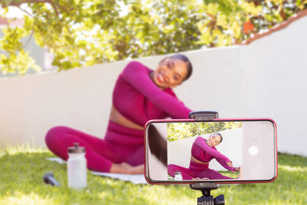 Smartphone-Bildschirm Aufnahme fröhlich attraktive sportliche schwarze Fitness-Bloggerin, die im Freien im Garten trainiert. Athletische afrikanisch-amerikanische Fitness-Trainerin streckt ihren Körper - Foto, Bild
