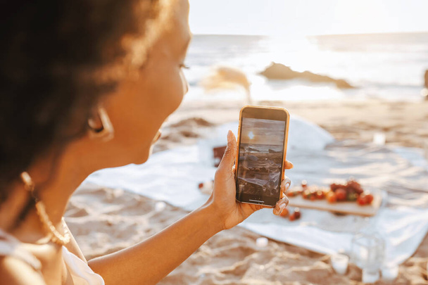 Mutlu kadın blogcu cep telefonu kullanıyor ve gün batımında plajda piknik yaparken fotoğraf çekiyor, telefona odaklanıyor. Açık hava eğlencesi, güneşli plajda dinlenme. - Fotoğraf, Görsel
