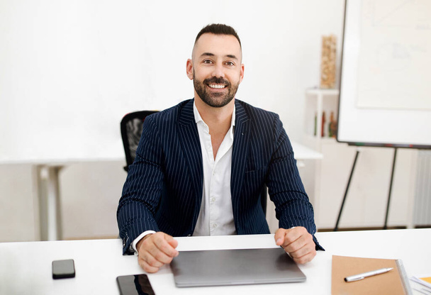 Hymyilevä valkoihoinen kypsä mies, jolla on parta puvussa, istuu pöydässä kannettavalla tietokoneella nykyaikaisessa coworking office sisustuksessa. Liikemies ja liike-elämä, opettaja etänä, johtaja työskentelee laitteen kanssa - Valokuva, kuva