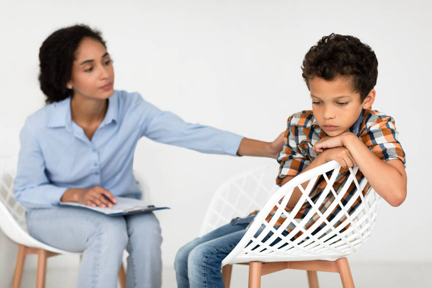 Depresión infantil. La psicóloga escolar Lady reconforta a un niño infeliz durante la sesión de psicoterapia sentado en interiores. Colegial deprimido visitando a un psicoterapeuta con problemas mentales. Enfoque selectivo - Foto, Imagen
