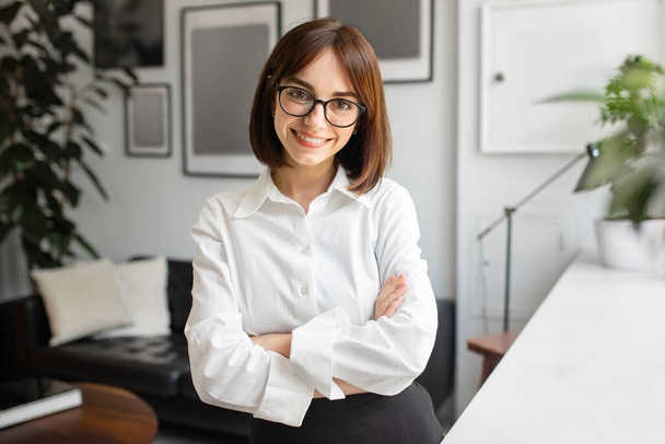 Başarılı bir girişimcilikti. Kameraya gülümseyen gözlüklü mutlu genç iş kadını portresi ofis içinde kollarını kavuşturmuş duruyor. İş kariyeri, istihdam kavramı - Fotoğraf, Görsel